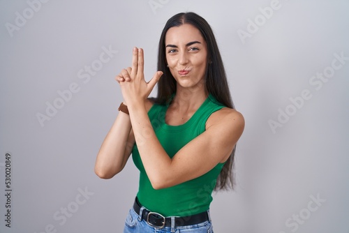Young woman standing over isolated background holding symbolic gun with hand gesture, playing killing shooting weapons, angry face