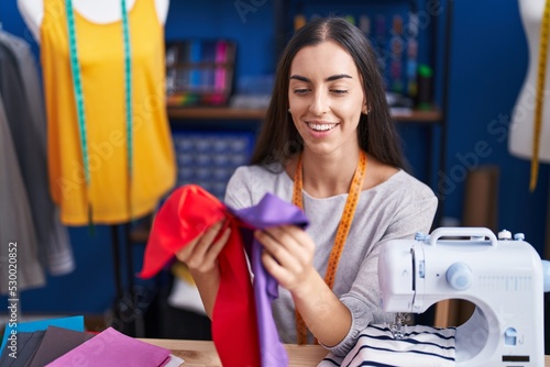 Young beautiful hispanic woman tailor smiling confident holding cloths at clothing factory