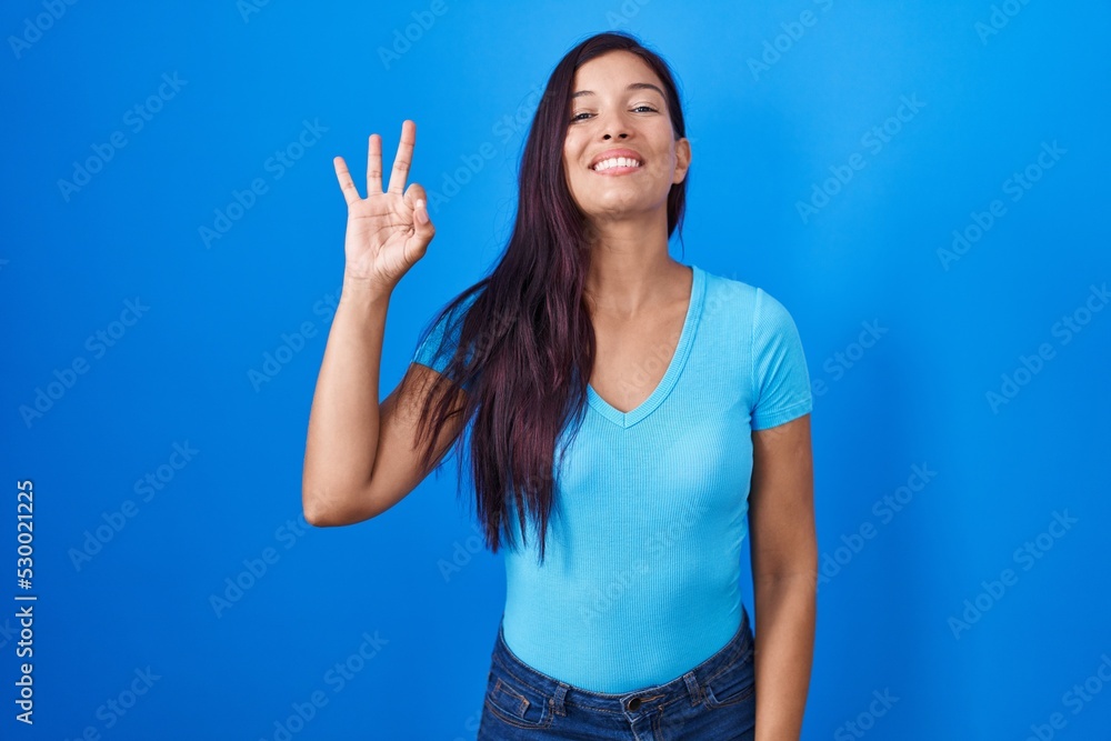 Fototapeta premium Young hispanic woman standing over blue background smiling positive doing ok sign with hand and fingers. successful expression.