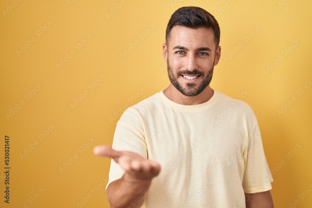 Handsome hispanic man standing over yellow background smiling cheerful offering palm hand giving assistance and acceptance. Handsome hispanic man standing over yellow background smiling cheerful offering palm hand giving assistance and acceptance.