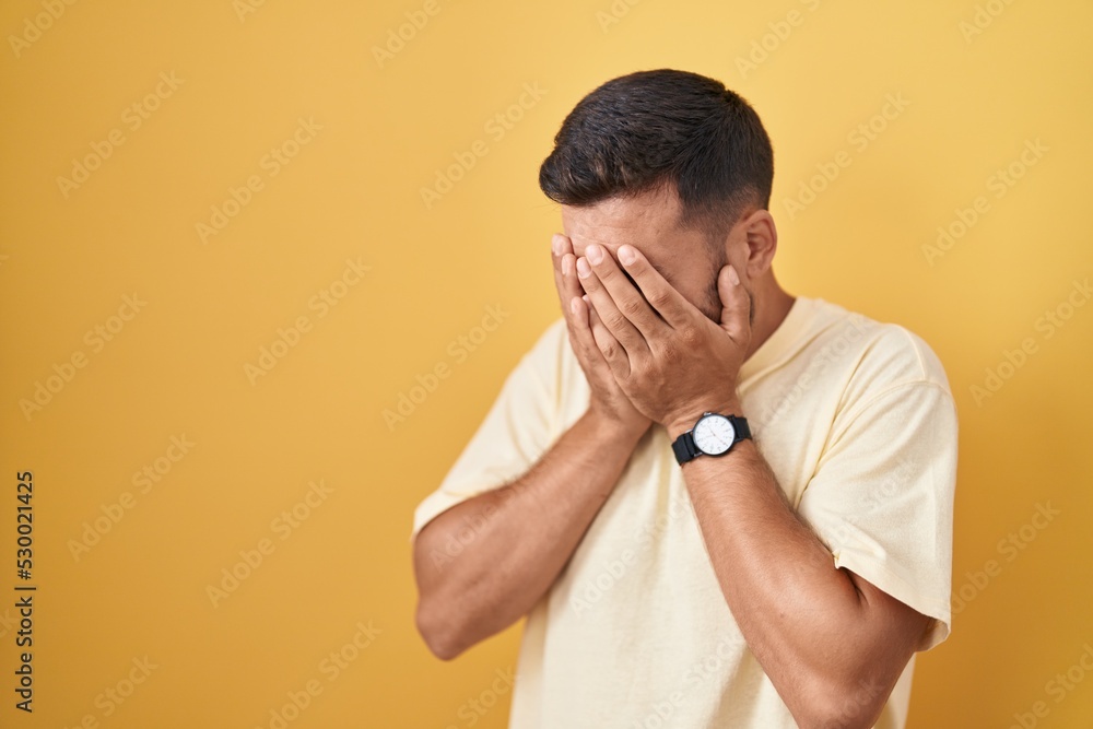 Handsome hispanic man standing over yellow background with sad expression covering face with hands while crying. depression concept. Handsome hispanic man standing over yellow background with sad expression covering face with hands while crying. depression concept.