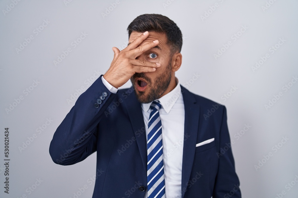 Handsome hispanic man wearing suit and tie peeking in shock covering face and eyes with hand, looking through fingers with embarrassed expression. Handsome hispanic man wearing suit and tie peeking in shock covering face and eyes with hand, looking through fingers with embarrassed expression.