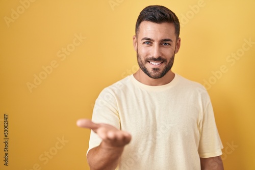 Handsome hispanic man standing over yellow background smiling cheerful offering palm hand giving assistance and acceptance.