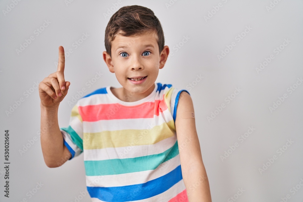 Young caucasian kid getting vaccine showing arm with band aid smiling with an idea or question pointing finger with happy face, number one Young caucasian kid getting vaccine showing arm with band aid smiling with an idea or question pointing finger with happy face, number one