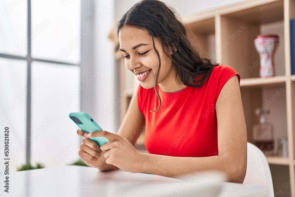 Young african american woman using smartphone sitting on table at home Young african american woman using smartphone sitting on table at home