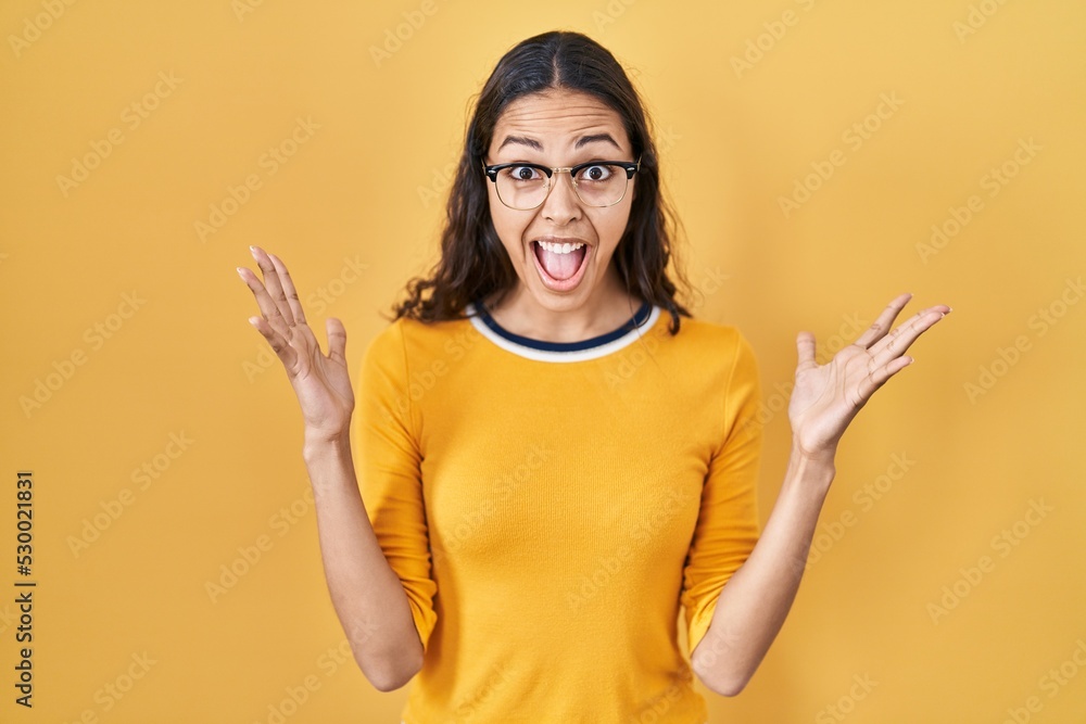 Young brazilian woman wearing glasses over yellow background celebrating victory with happy smile and winner expression with raised hands Young brazilian woman wearing glasses over yellow background celebrating victory with happy smile and winner expression with raised hands
