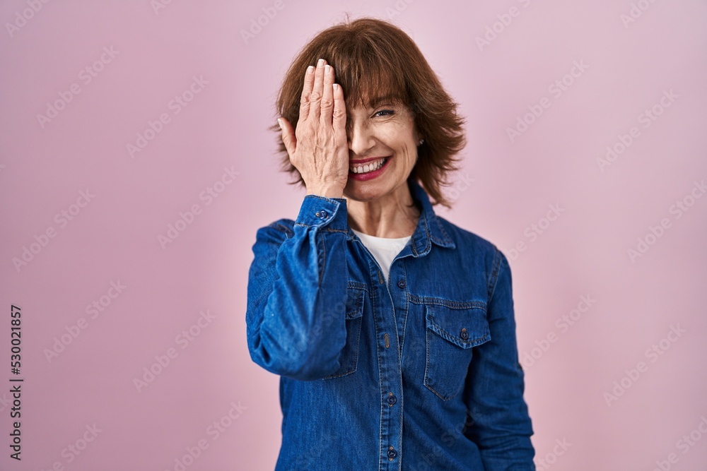 Middle age woman standing over pink background covering one eye with hand, confident smile on face and surprise emotion.
