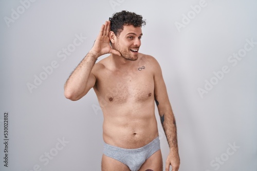 Young hispanic man standing shirtless wearing underware smiling with hand over ear listening an hearing to rumor or gossip. deafness concept.