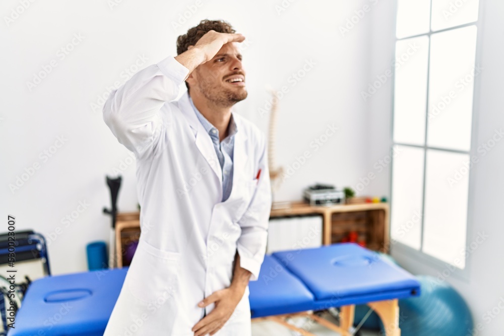 Handsome young man working at pain recovery clinic very happy and smiling looking far away with hand over head. searching concept. Handsome young man working at pain recovery clinic very happy and smiling looking far away with hand over head. searching concept.