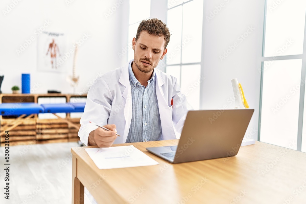 Young hispanic man wearing physiotherapist uniform writing on document at clinic