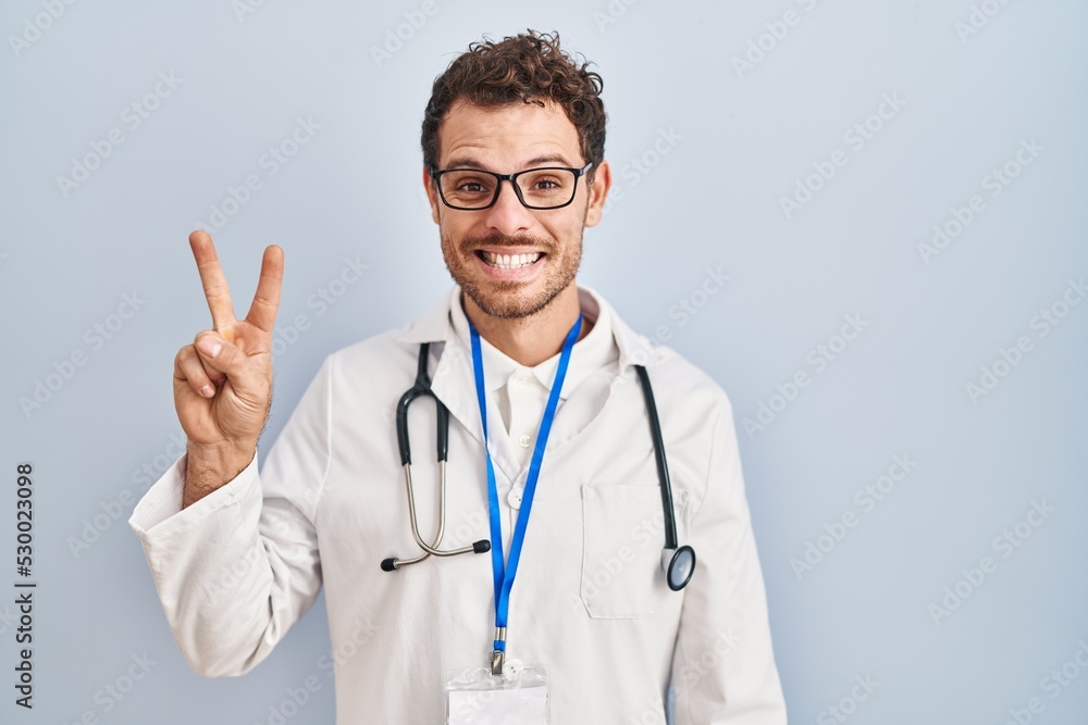 Young hispanic man wearing doctor uniform and stethoscope showing and pointing up with fingers number two while smiling confident and happy. Young hispanic man wearing doctor uniform and stethoscope showing and pointing up with fingers number two while smiling confident and happy.