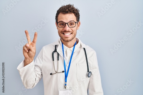 Young hispanic man wearing doctor uniform and stethoscope showing and pointing up with fingers number two while smiling confident and happy.