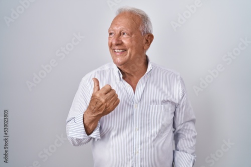 Senior man with grey hair standing over isolated background smiling with happy face looking and pointing to the side with thumb up. © Krakenimages.com