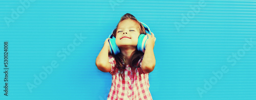 Portrait of happy little girl child in wireless headphones listening to music on blue background, blank copy space for advertising text