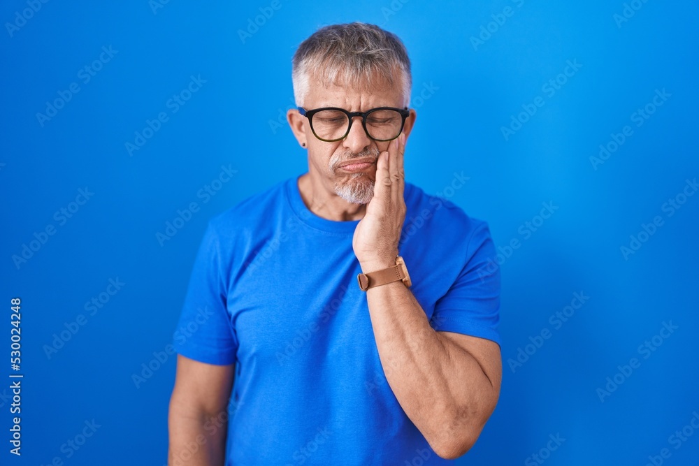 Hispanic man with grey hair standing over blue background touching mouth with hand with painful expression because of toothache or dental illness on teeth. dentist Hispanic man with grey hair standing over blue background touching mouth with hand with painful expression because of toothache or dental illness on teeth. dentist