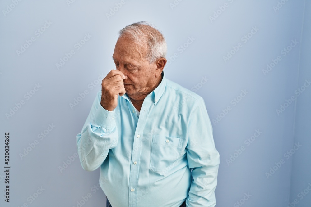 Senior man with grey hair standing over blue background tired rubbing nose and eyes feeling fatigue and headache. stress and frustration concept.