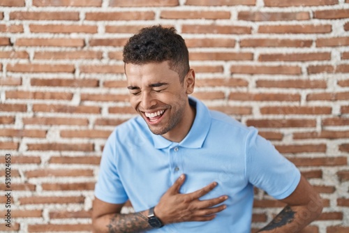 Brazilian young man standing over brick wall smiling and laughing hard out loud because funny crazy joke with hands on body.