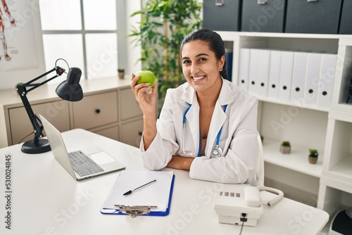Young hispanic woman wearing dietitian uniform holding apple at clinic