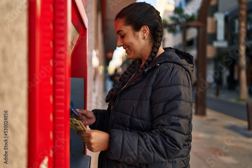 фотографія Young hispanic woman holding money of automatic teller bank machine at street