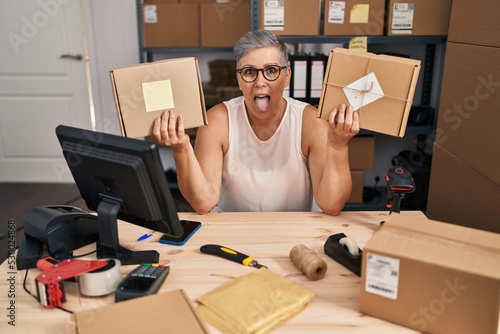 Middle age woman working at small business ecommerce sticking tongue out happy with funny expression.