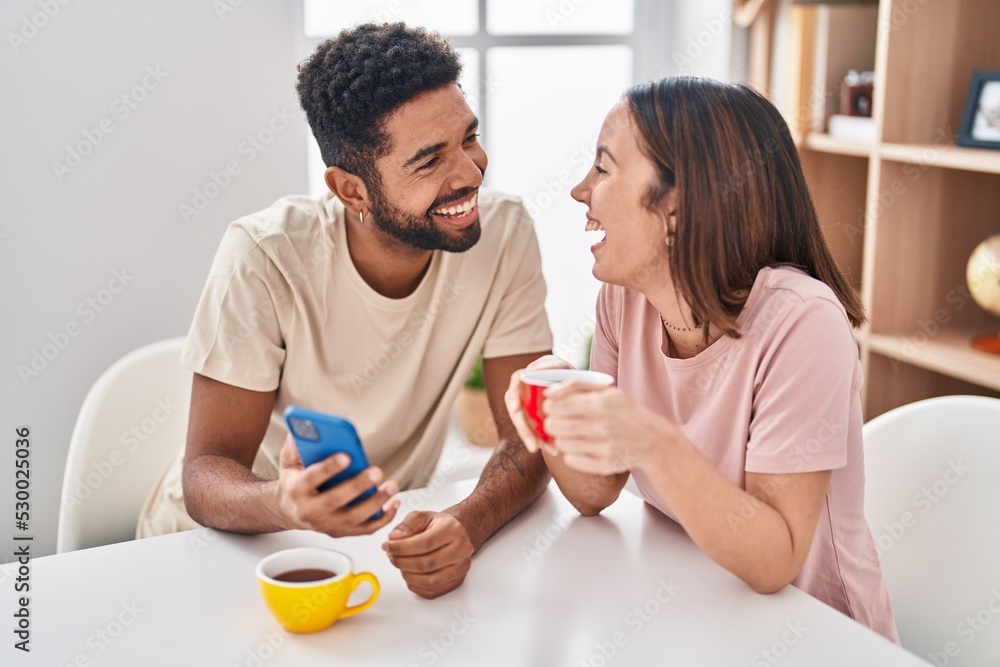 Man and woman couple sitting on table drinking coffee using smartphone at home Man and woman couple sitting on table drinking coffee using smartphone at home