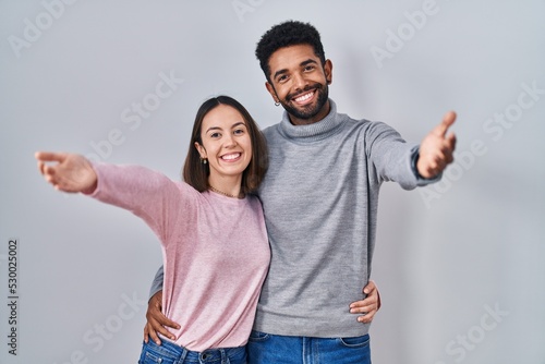Young hispanic couple standing together looking at the camera smiling with open arms for hug. cheerful expression embracing happiness.