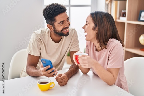 Man and woman couple sitting on table drinking coffee using smartphone at home
