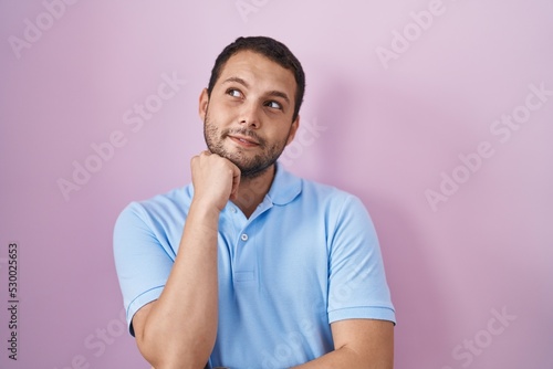 Hispanic man standing over pink background with hand on chin thinking about question, pensive expression. smiling with thoughtful face. doubt concept.