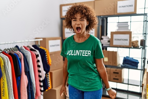 Young african american woman wearing volunteer t shirt at donations stand angry and mad screaming frustrated and furious, shouting with anger. rage and aggressive concept.