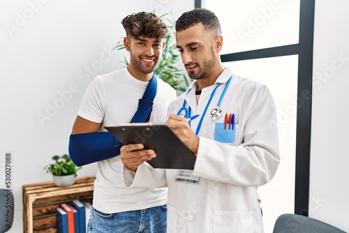 Two hispanic men doctor and patient writing on clipboard standing at hospital waiting room