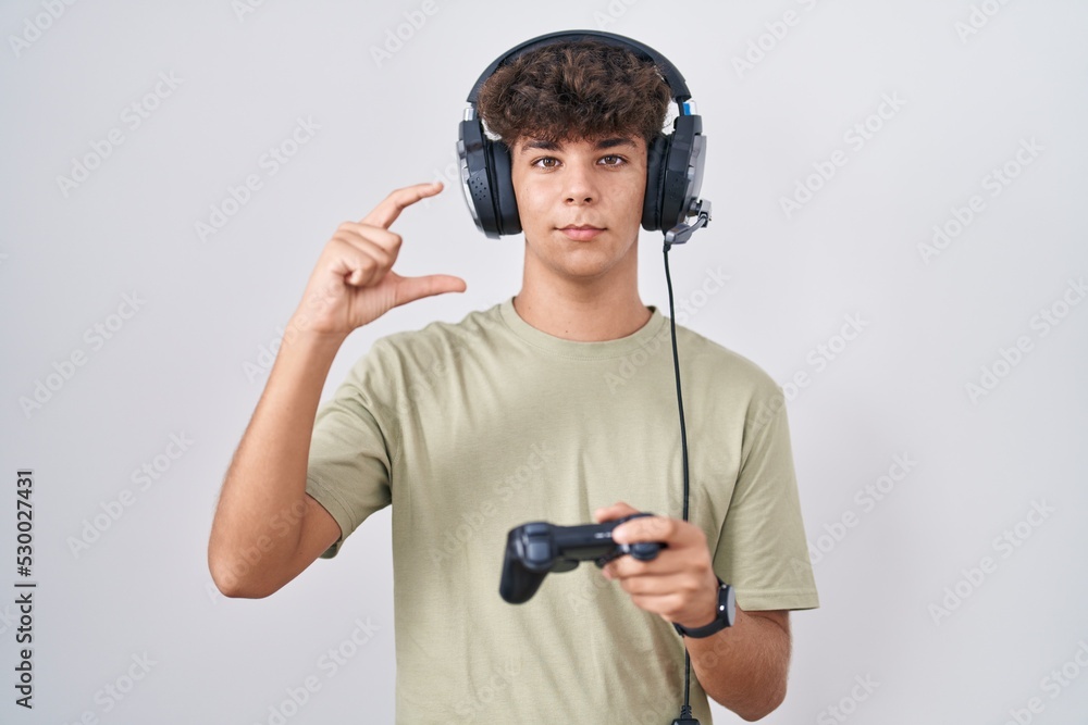 Hispanic teenager playing video game holding controller smiling and confident gesturing with hand doing small size sign with fingers looking and the camera. measure concept. Hispanic teenager playing video game holding controller smiling and confident gesturing with hand doing small size sign with fingers looking and the camera. measure concept.