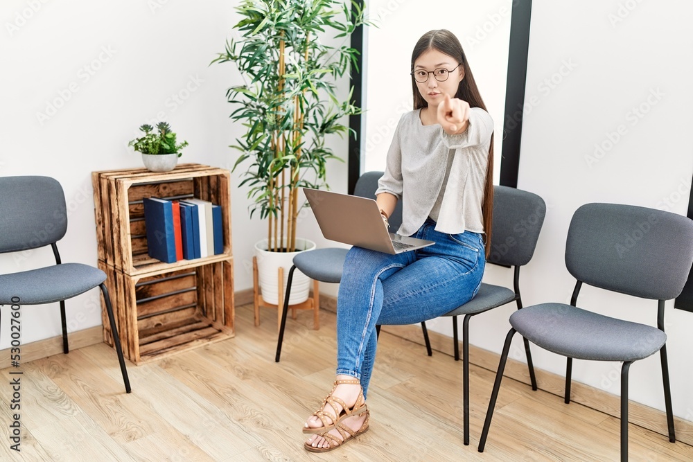 Young asian nurse woman sitting at waiting room using laptop pointing with finger to the camera and to you, confident gesture looking serious Young asian nurse woman sitting at waiting room using laptop pointing with finger to the camera and to you, confident gesture looking serious