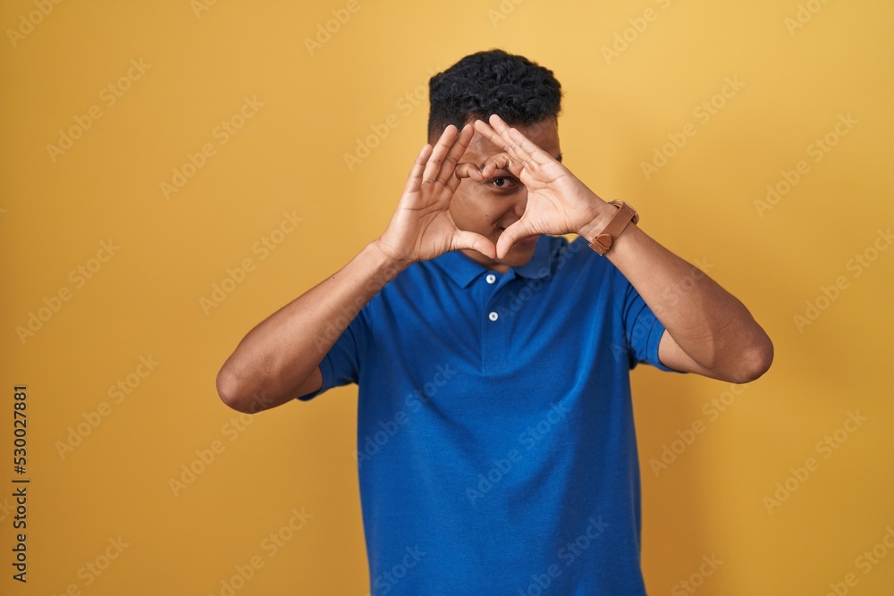 Young hispanic man standing over yellow background doing heart shape with hand and fingers smiling looking through sign Young hispanic man standing over yellow background doing heart shape with hand and fingers smiling looking through sign