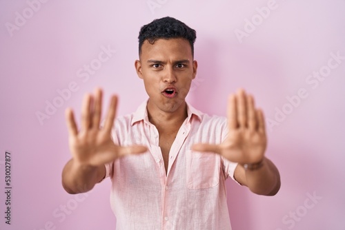 Young hispanic man standing over pink background doing stop gesture with hands palms, angry and frustration expression