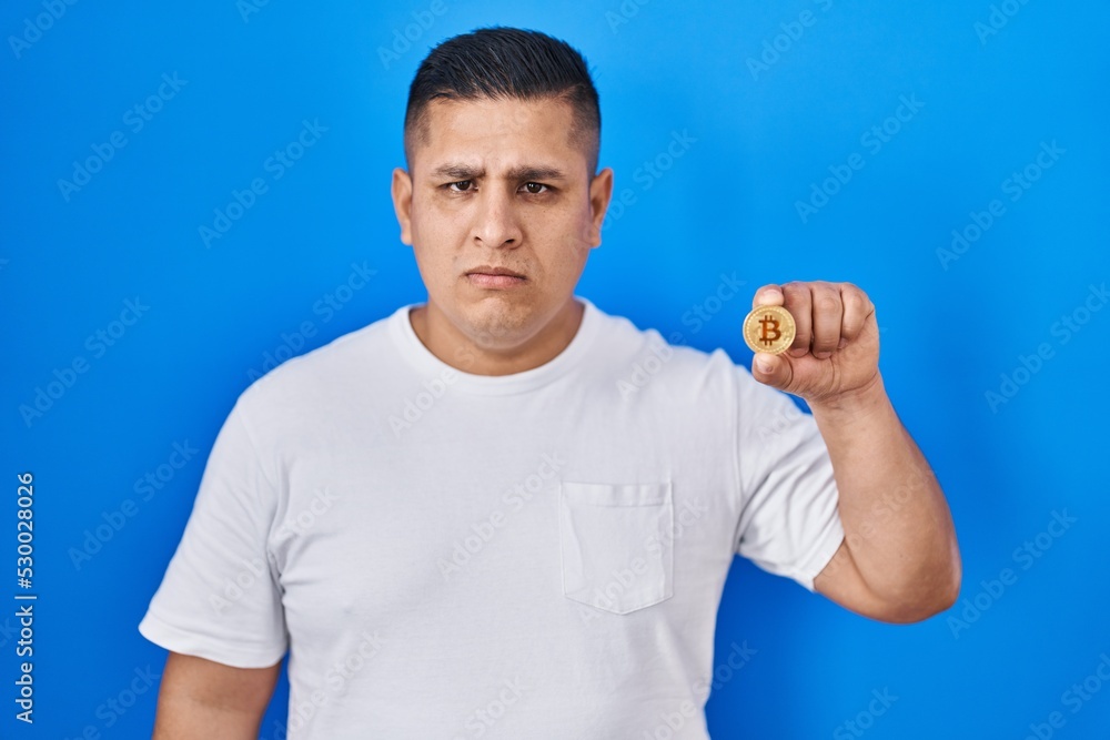 Hispanic young man holding virtual currency bitcoin thinking attitude and sober expression looking self confident Hispanic young man holding virtual currency bitcoin thinking attitude and sober expression looking self confident