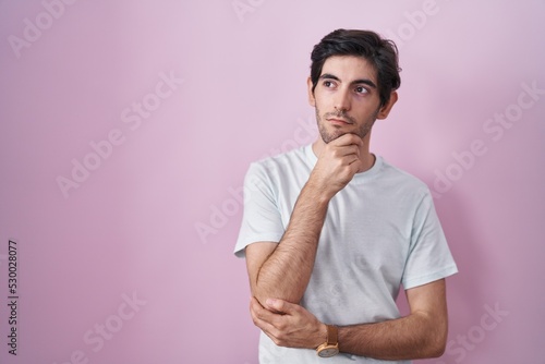 Young hispanic man standing over pink background with hand on chin thinking about question, pensive expression. smiling with thoughtful face. doubt concept.