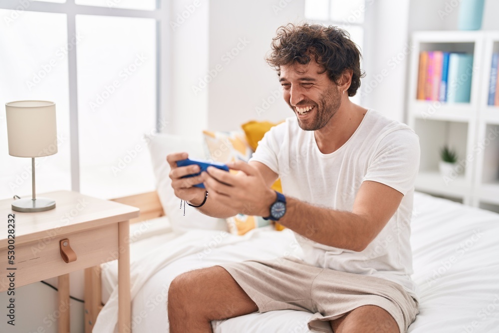 Young hispanic man watching video on smartphone sitting on bed at bedroom Young hispanic man watching video on smartphone sitting on bed at bedroom