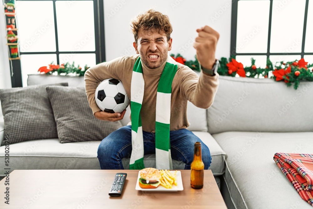 Young hispanic man football hooligan holding ball at home annoyed and frustrated shouting with anger, yelling crazy with anger and hand raised Young hispanic man football hooligan holding ball at home annoyed and frustrated shouting with anger, yelling crazy with anger and hand raised