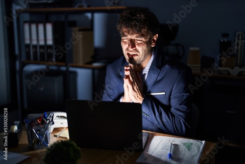 Hispanic young man working at the office at night begging and praying with hands together with hope expression on face very emotional and worried. begging.