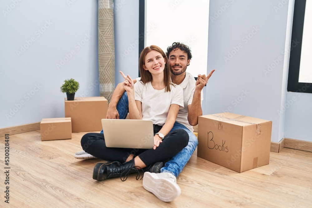 Young couple using laptop at new home with a big smile on face, pointing with hand finger to the side looking at the camera. Young couple using laptop at new home with a big smile on face, pointing with hand finger to the side looking at the camera.