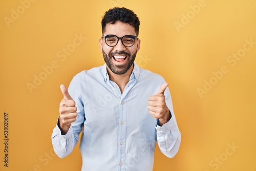 Hispanic man with beard standing over yellow background success sign doing positive gesture with hand, thumbs up smiling and happy. cheerful expression and winner gesture. © Krakenimages.com