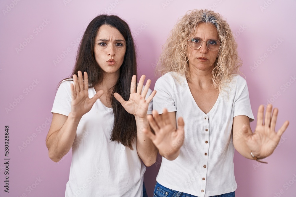 Mother and daughter standing together over pink background moving away hands palms showing refusal and denial with afraid and disgusting expression. stop and forbidden. Mother and daughter standing together over pink background moving away hands palms showing refusal and denial with afraid and disgusting expression. stop and forbidden.
