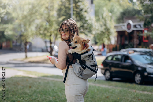 Happy smiling woman walking outdoors in city parkland with dog Welsh Corgi Pembroke in a special backpack © bondvit