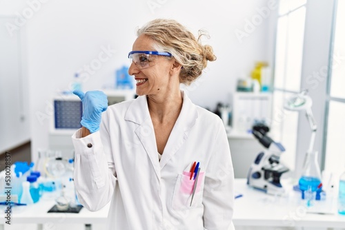 Middle age blonde woman working at scientist laboratory smiling with happy face looking and pointing to the side with thumb up.
