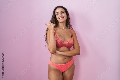 Young hispanic woman wearing lingerie over pink background smiling with happy face looking and pointing to the side with thumb up.