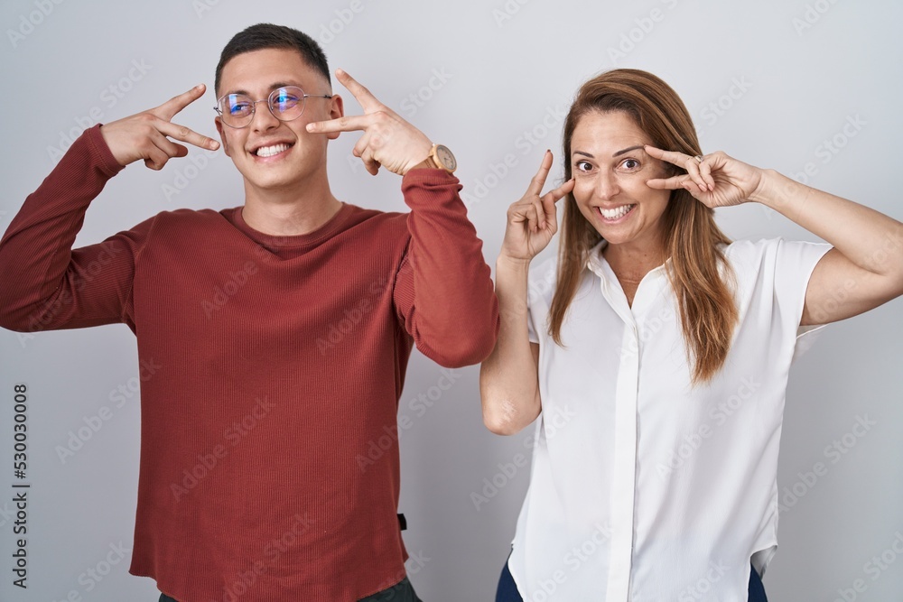 Mother and son standing together over isolated background doing peace symbol with fingers over face, smiling cheerful showing victory Mother and son standing together over isolated background doing peace symbol with fingers over face, smiling cheerful showing victory