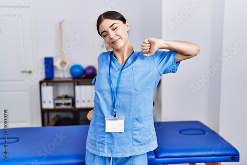 Young hispanic woman wearing physiotherapist uniform standing at clinic stretching back  tired and relaxed  sleepy and yawning for early morning