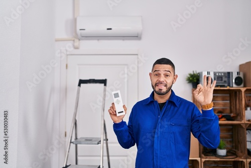 Hispanic repairman working with air conditioner doing ok sign with fingers, smiling friendly gesturing excellent symbol