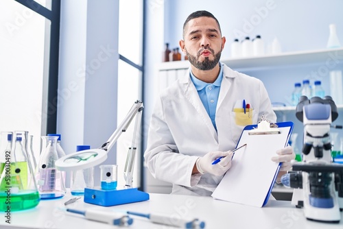Hispanic man working at scientist laboratory holding blank clipboard looking at the camera blowing a kiss being lovely and sexy. love expression.