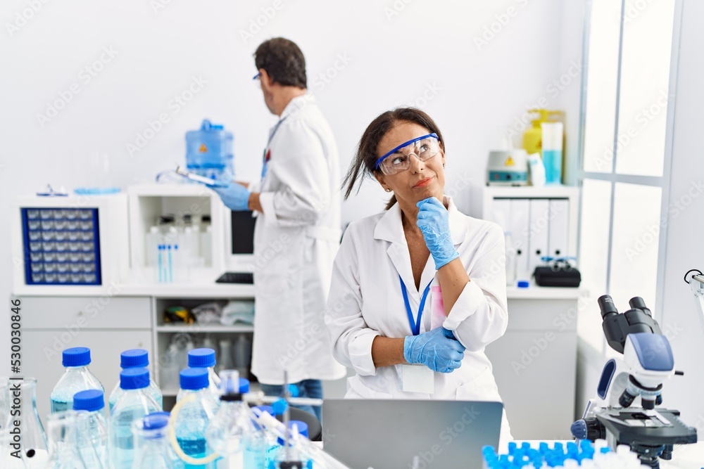 Middle age woman working at scientist laboratory serious face thinking about question with hand on chin, thoughtful about confusing idea Middle age woman working at scientist laboratory serious face thinking about question with hand on chin, thoughtful about confusing idea